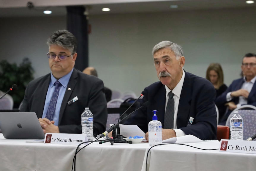 Two men sit at a desk at a parliamentary inquiry.