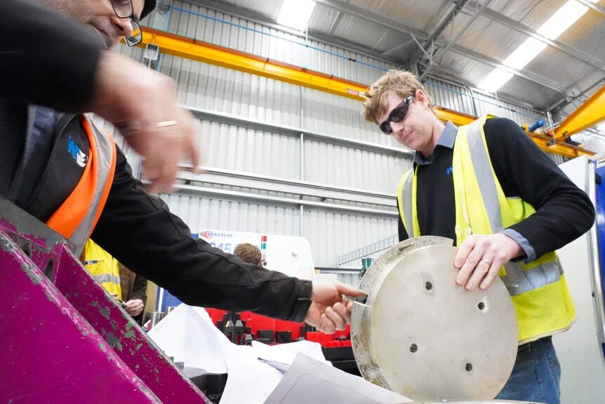 A man in safety glasses and a high vis vest holds a metal mould in a workshop.