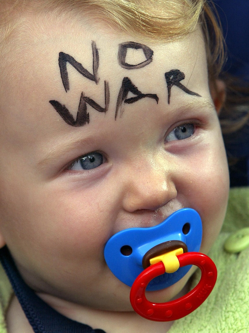 A happy baby with a dummy in its mouth has no-war painted on it's forehead.
