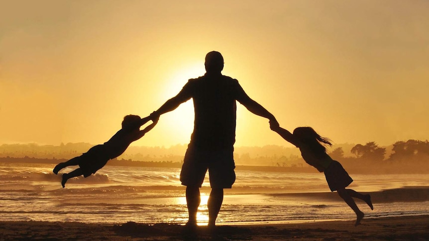 A silhouette of two young children swinging off their dad's arms on the beach.