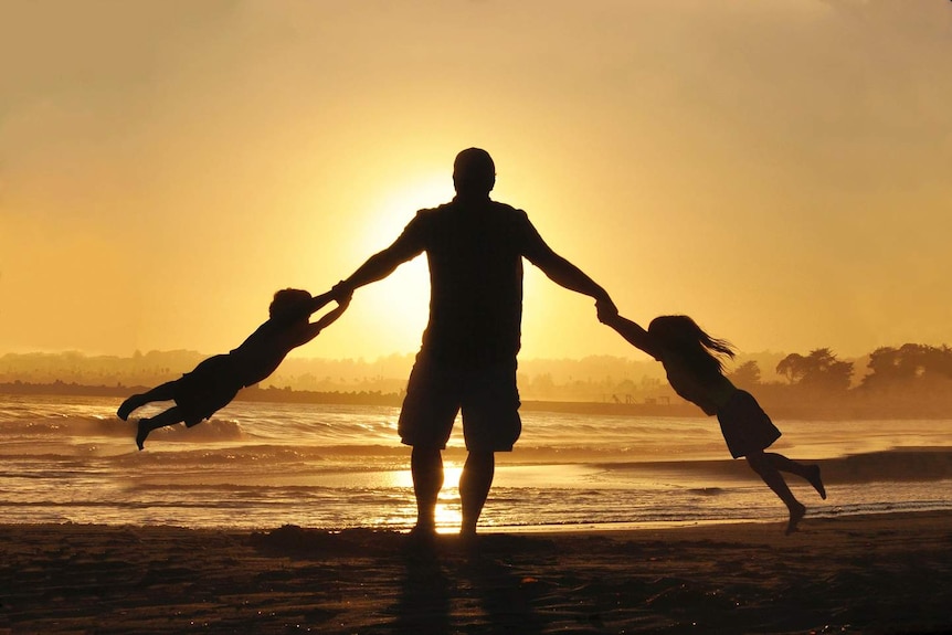 A silhouette of two young children swinging off their dad's arms on the beach.