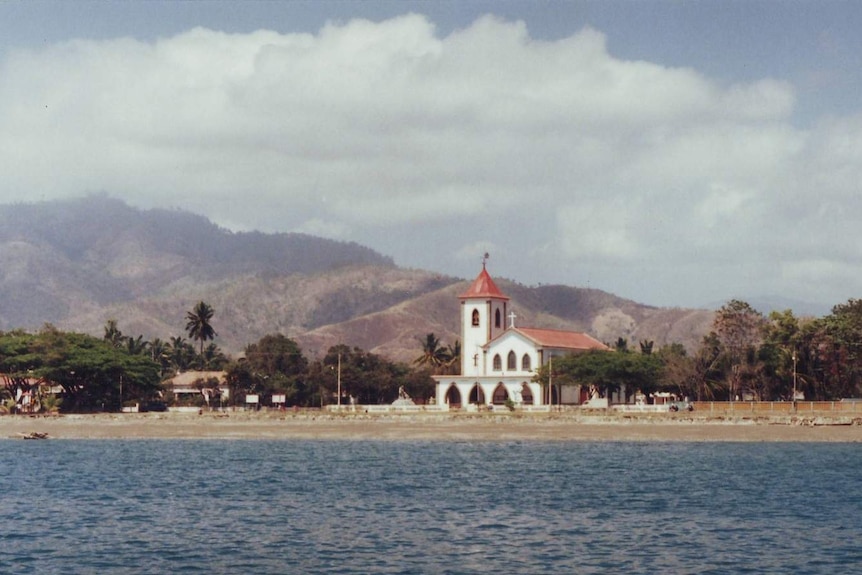 A view from a boat looks across blue water to a red-roofed white Spanish-style church with mountains in the distance.