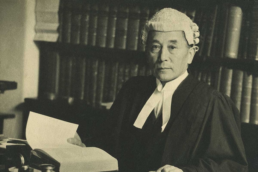 Man dressed in old barrister attire, and a wig, sits at his desk.