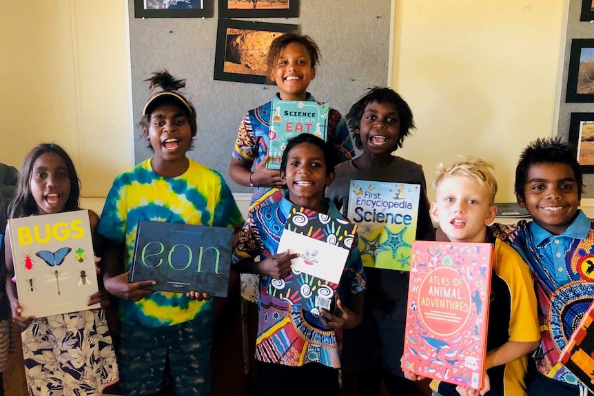 Children smile and hold books as they look into the camera for a photo taken in a classroom.