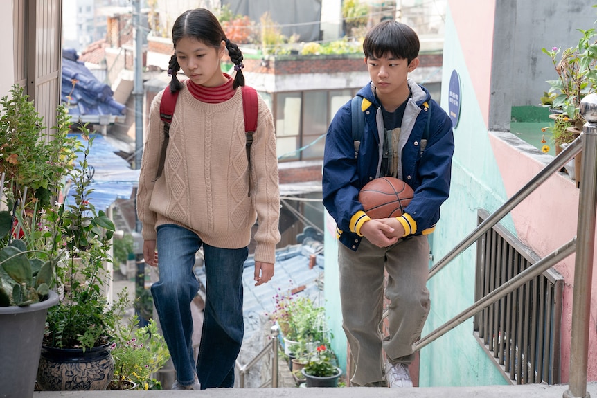 A young Korean girl in jeans a cream jumper walks up stairs beside a shy-looking Korean boy in blue jacket, holding a basketball
