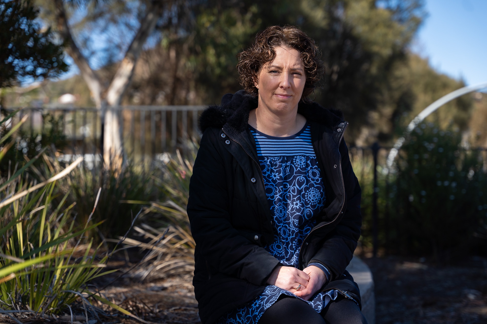 A woman with dark curly hair sits in the bush in a park.