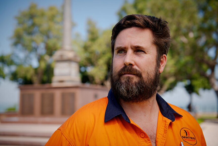 A man in an orange work shirt looks off camera with a serious expression. Behind him is the Darwin Cenotaph.