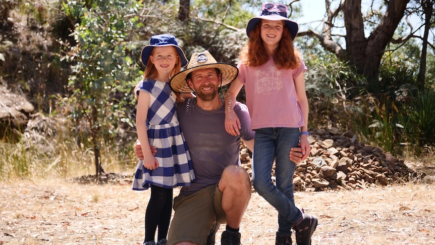 A man crouches and smiles with his two daughters amongst trees.
