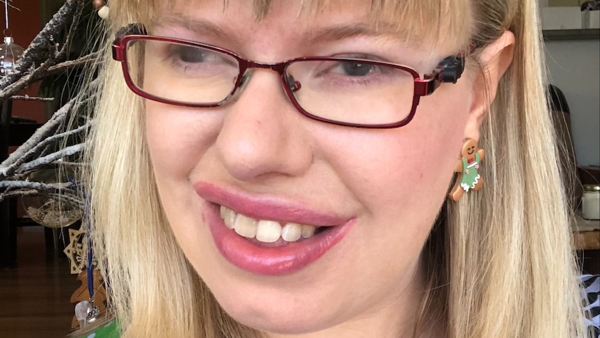 A smiling young woman with a blonde bob and fringe wears red framed glasses, festive earrings and blue patterned top
