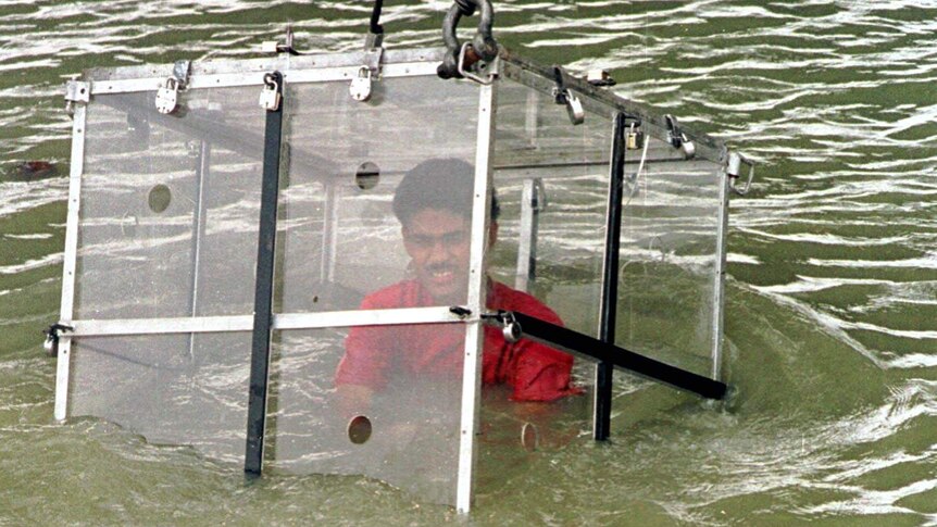 A crane lowers a man sitting inside a locked glass box into a river.