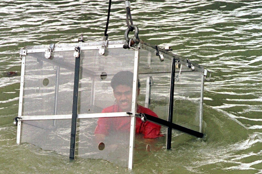 A crane lowers a man sitting inside a locked glass box into a river.