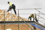 Three builders stand on the roof and scaffolding of a half-built brick home.
