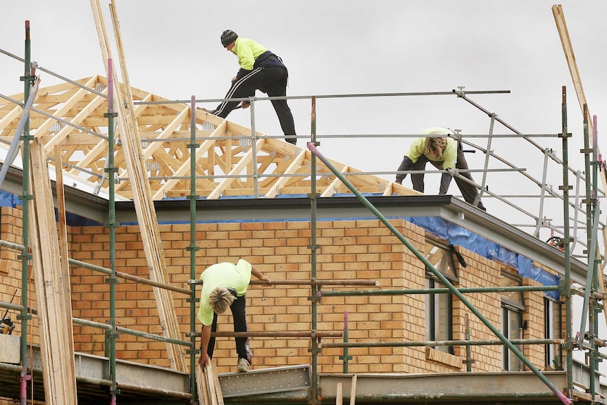 Three builders stand on the roof and scaffolding of a half-built brick home.