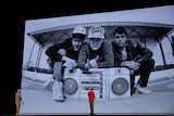 Two men stand on stage in front of large screen with black and white photo of hip hop group Beastie Boys crouching with boombox.