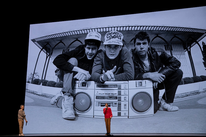 Two men stand on stage in front of large screen with black and white photo of hip hop group Beastie Boys crouching with boombox.