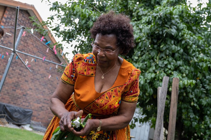Theresa leans over picking green herbs from her garden in an orange dress.