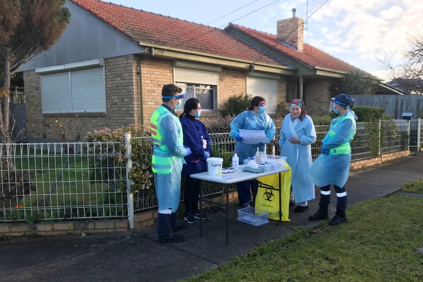 Four health workers wearing PPE speak to a woman in a dressing gown at a coronavirus testing station on a residential street.