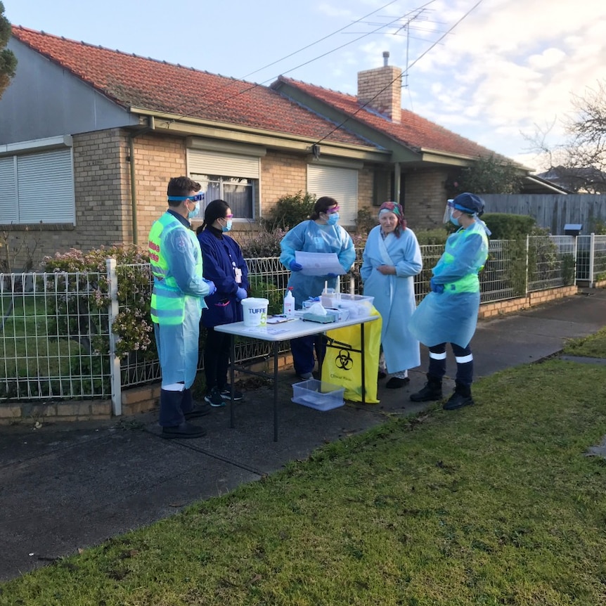Four health workers wearing PPE speak to a woman in a dressing gown at a coronavirus testing station on a residential street.