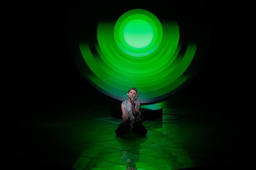 A female singer crouches on stage, singing her Eurovision song, while behind her is a large green glowing disc.
