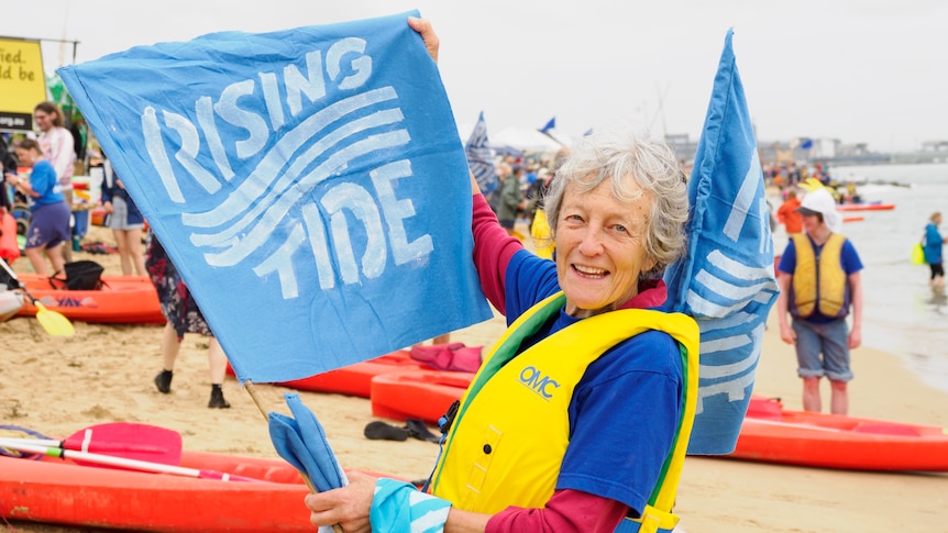 An older lady wearing a life jacket holds a blue flag that reads, RISING TIDE.