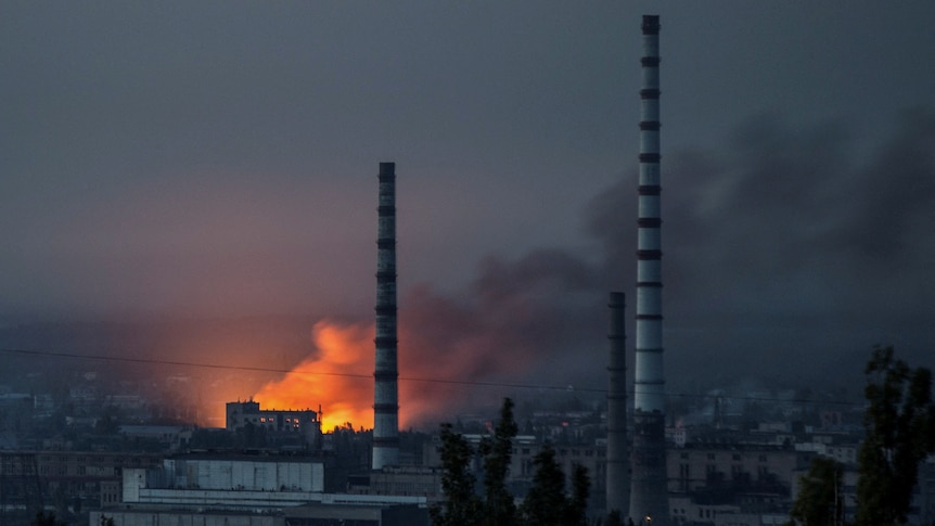 Smoke and flame rise from a building, with two large thin exhaust towers reaching into the sky in the foreground.