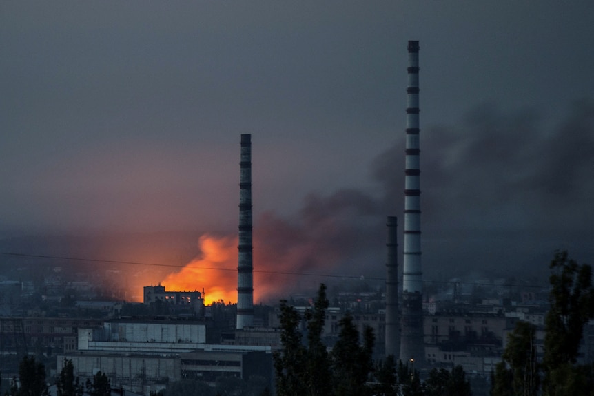 Smoke and flame rise from a building, with two large thin exhaust towers reaching into the sky in the foreground.