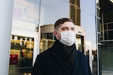 A white man in a dark coat standing outside a large glass building and wearing a face mask