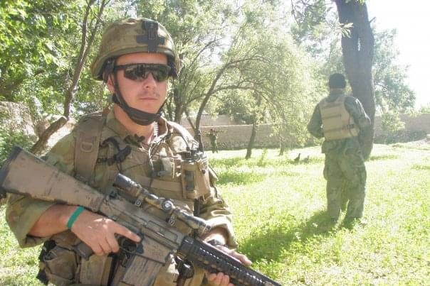 Phil Thompson dressed in army camo stands holding gun with helmet and sunglasses