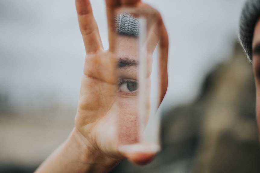 Woman holds a mirror and looks at reflection to depict the self-doubt and self esteem issues that arise from imposter syndrome.