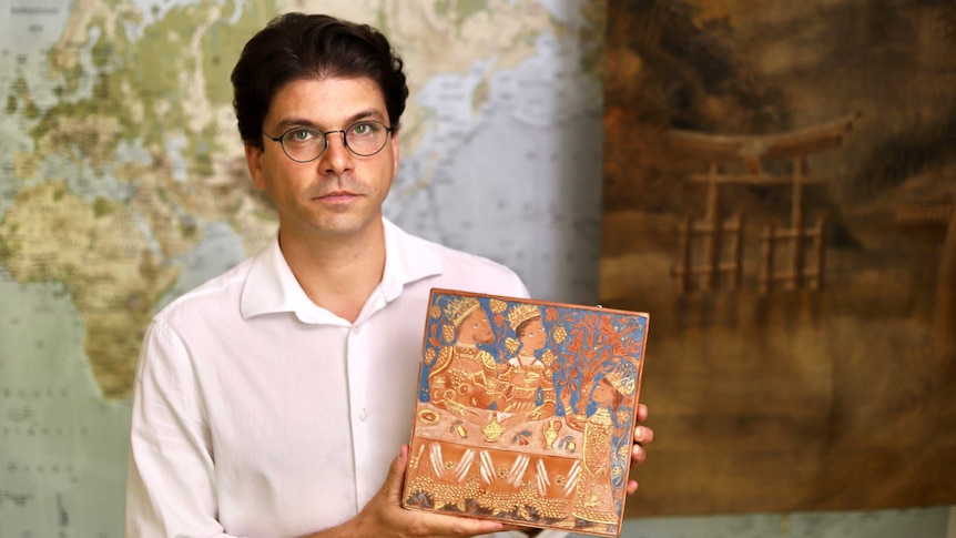 A man wearing glasses sits in front of a world map holding a tile with an ornate design on it.