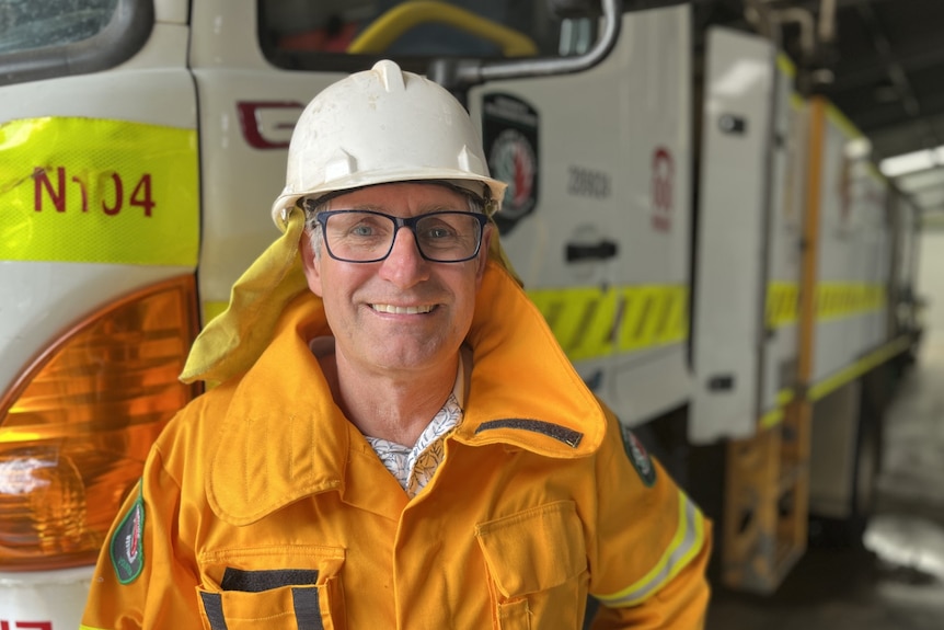 A smiling middle-ageg man in a yellow coat and hard hat in front of a  fire truck.