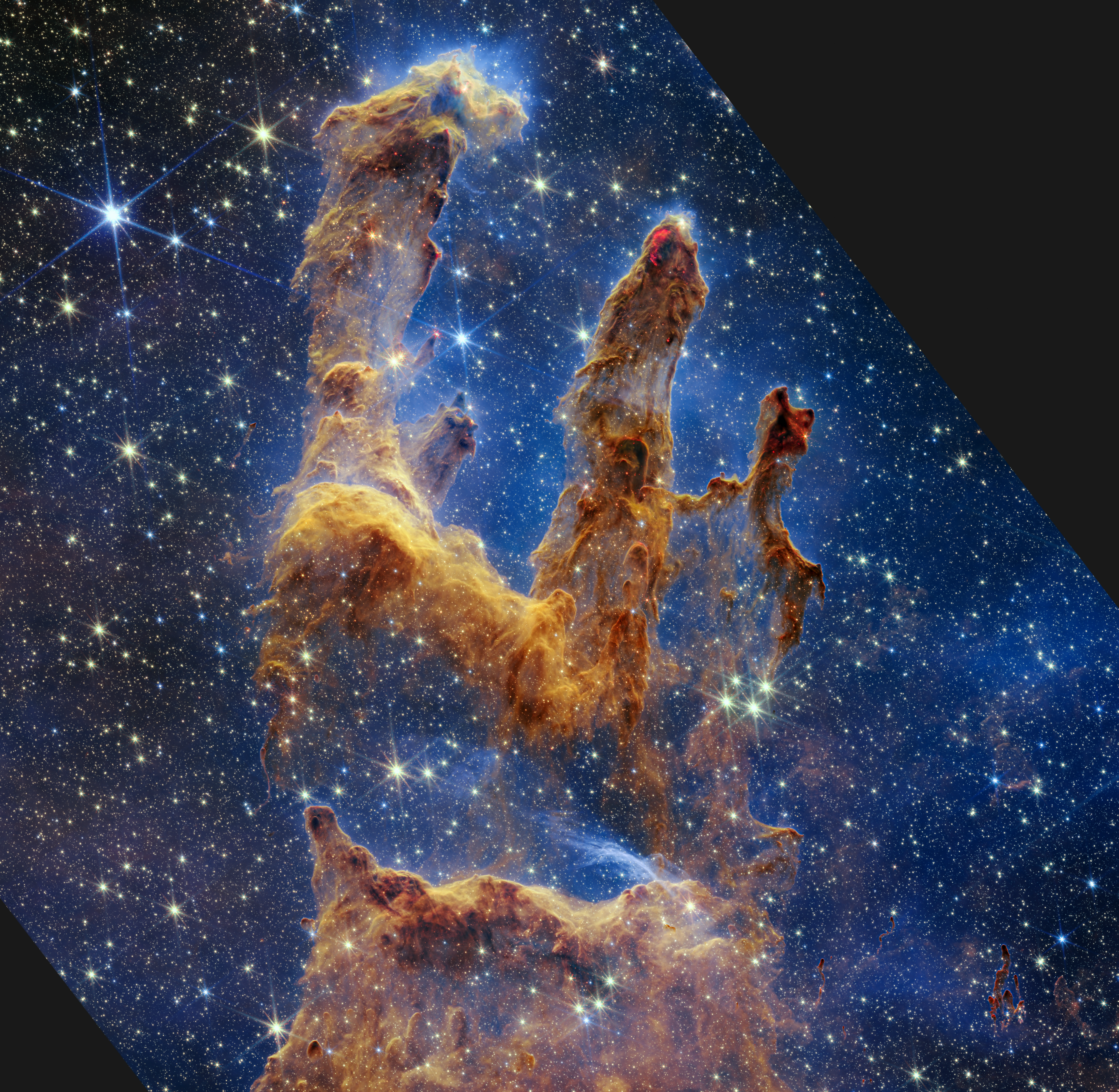 Imagen de los Pilares de la Creación, con nubes de polvo de color marrón anaranjado sobre un fondo azul oscuro estrellado. 