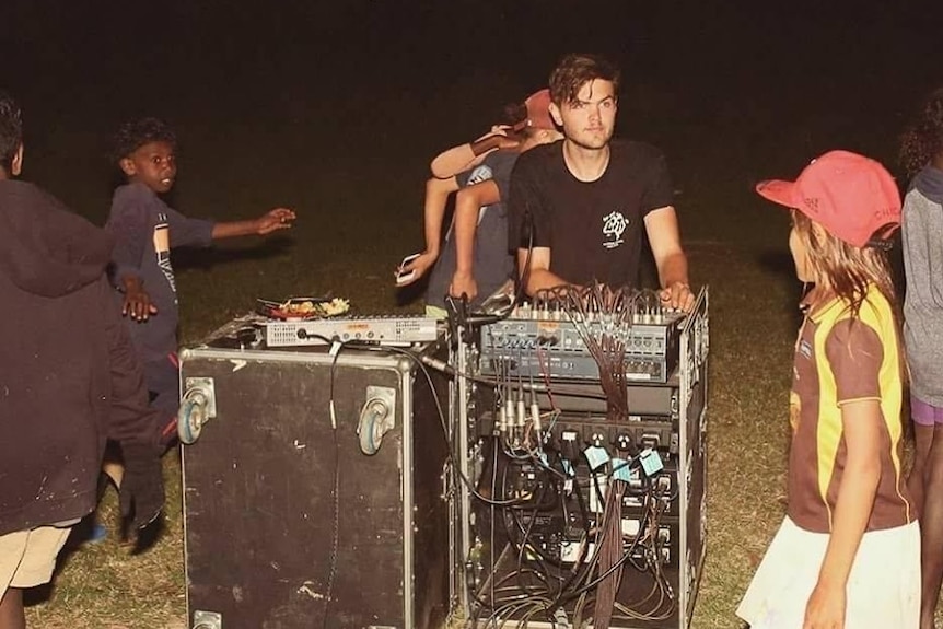 A sound engineer stands at a sound box at an outdoor gig in the evening.