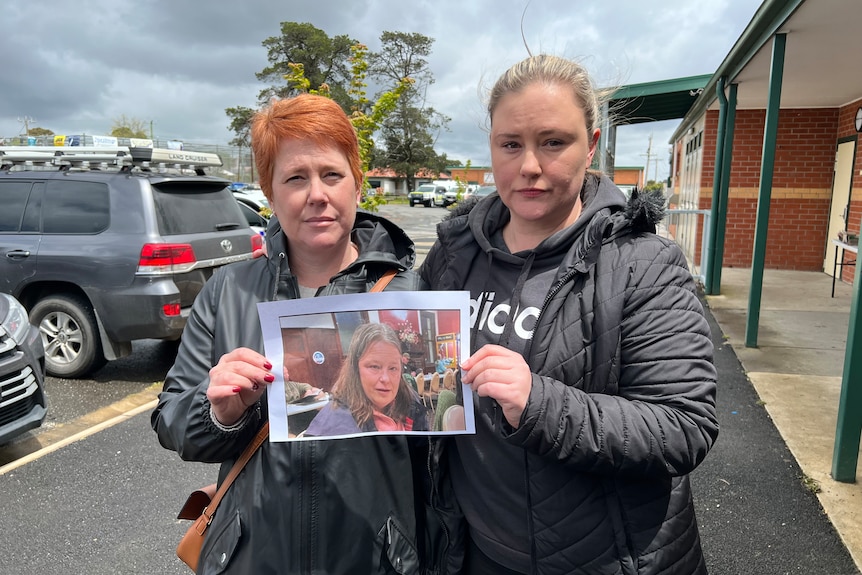 Two women dressed for warmth stand outside holding a picture of a missing woman.