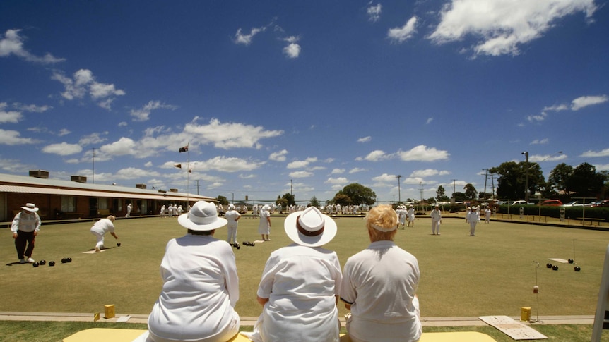 Three women sit on a bench watching games of lawn bowls in Brisbane.