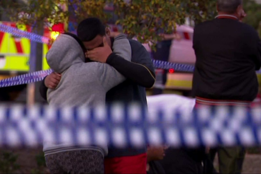 A man and a woman embrace each other and weep in an area surrounded by police tape.