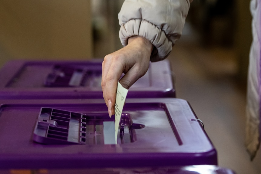 A hand with painted acrylic nails drops a ballot paper into a slot on top of a purple plastic box