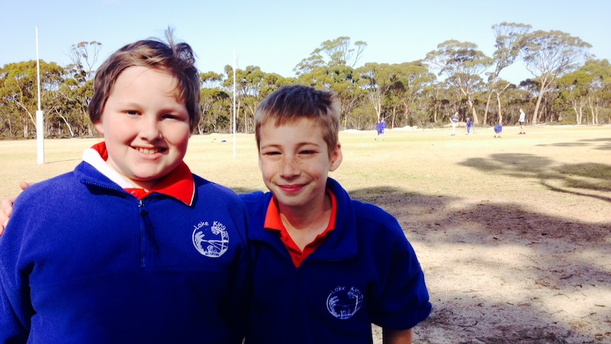 Two 11 year old boys, wearing blue and red school uniforms stand arm in arm in the shade of gum trees at a rural school oval