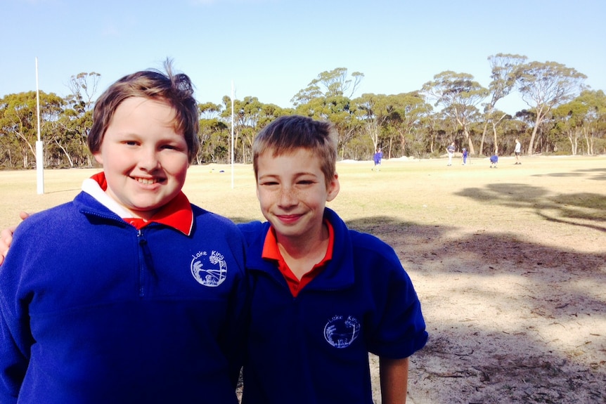 Two 11 year old boys, wearing blue and red school uniforms stand arm in arm in the shade of gum trees at a rural school oval