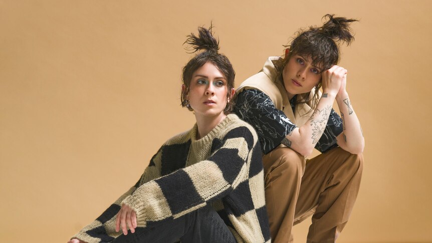 Twins Tegan and Sara sitting in front of a beige background, wearing baggy jumpers and high little ponytails