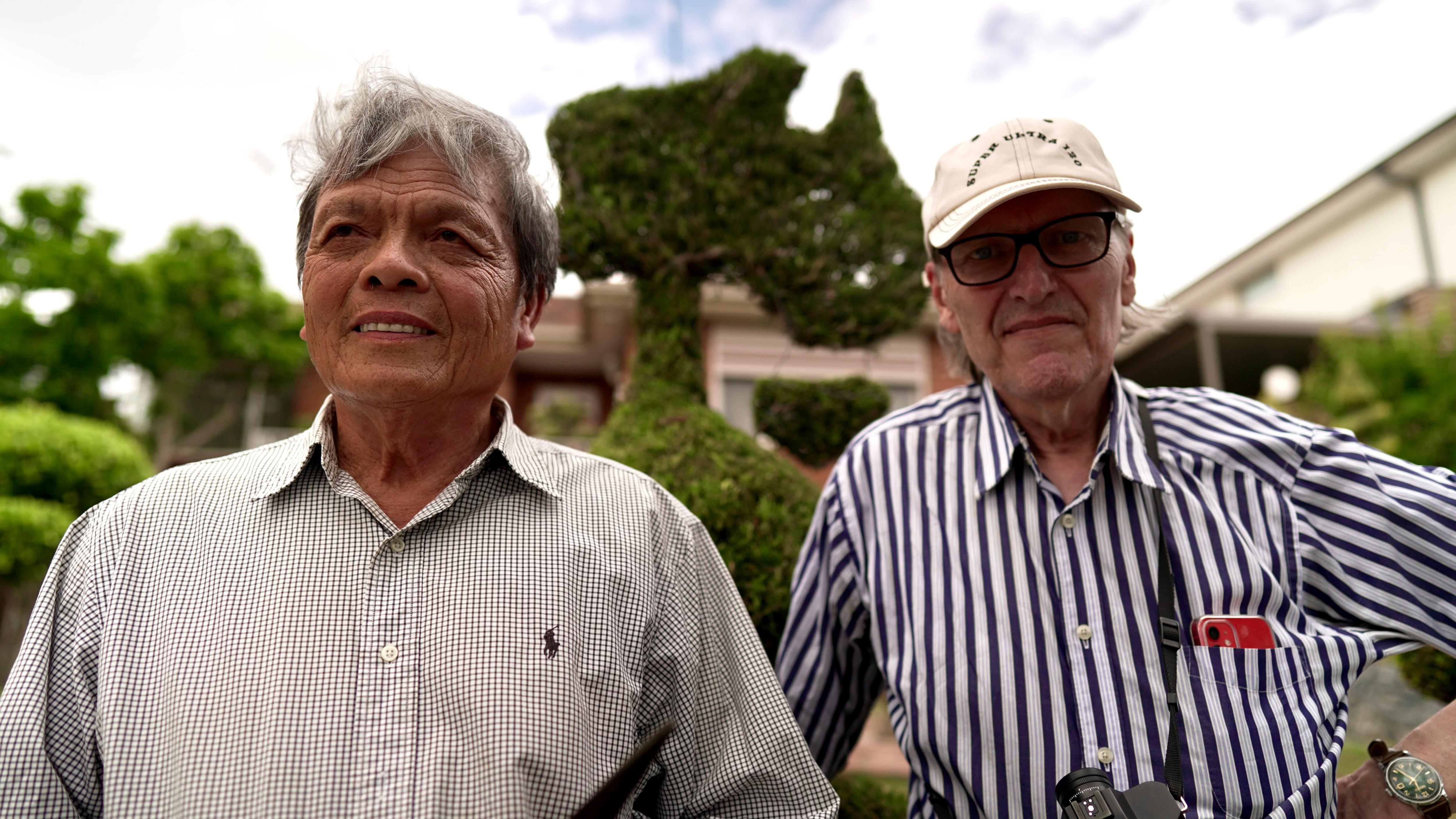 Two men are pictured standing next to each other, behind them is a hedge trimmed into an art piece.