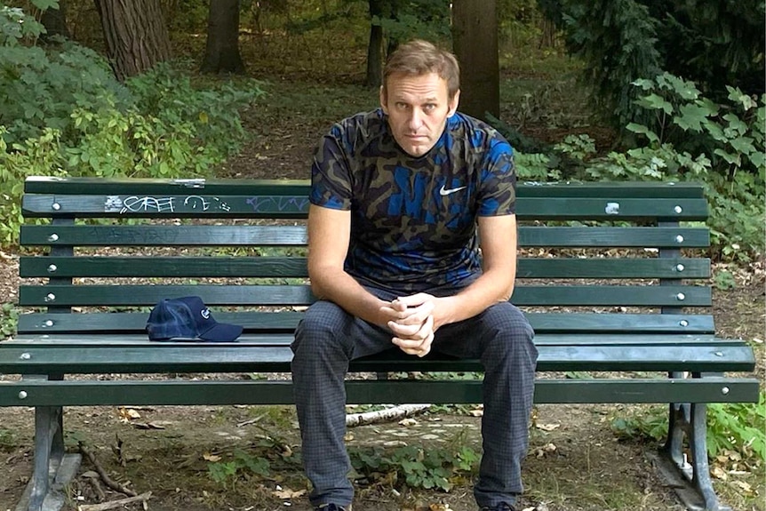 Navalny sits on a bench looking straight at camera with hands clasped and elbows on his knees. There are trees in the background