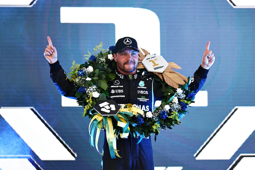 Valtteri Bottas wears a garland of flowers around his neck with a 1 on it, and holds his fingers up to the sky