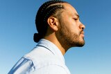 Side profile photo of Ziggy Ramo. The sky is cloudless blue and he's wearing a light blue button up shirt. His hair is braided.