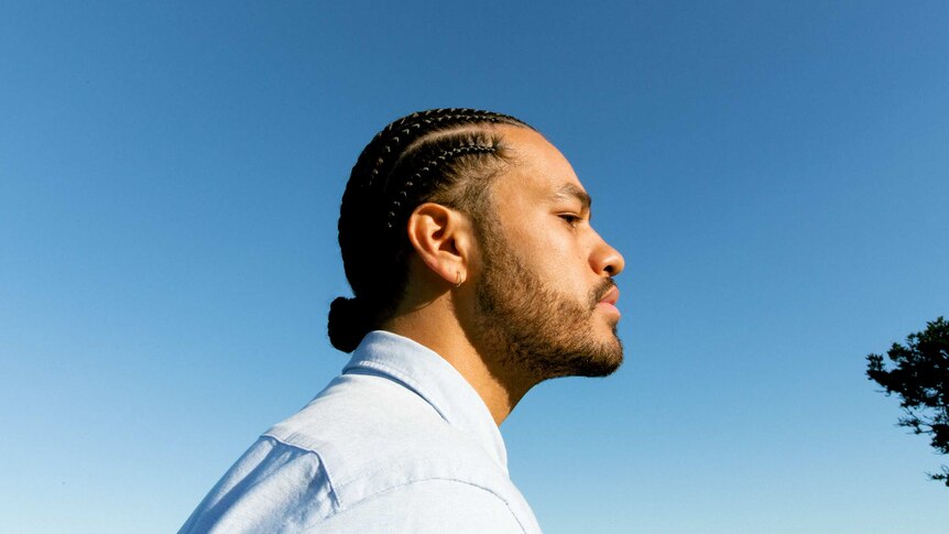 Side profile photo of Ziggy Ramo. The sky is cloudless blue and he's wearing a light blue button up shirt. His hair is braided.