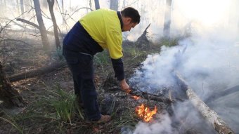 Peter Dixon looks at a fire he is stoking on the ground, as smoke billows on either side of him in a forest.