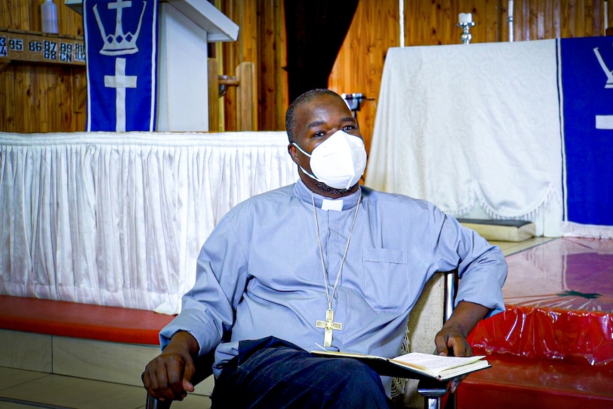 A pastor in a blue shirt and clerical collar with a cross necklace on sits in a church
