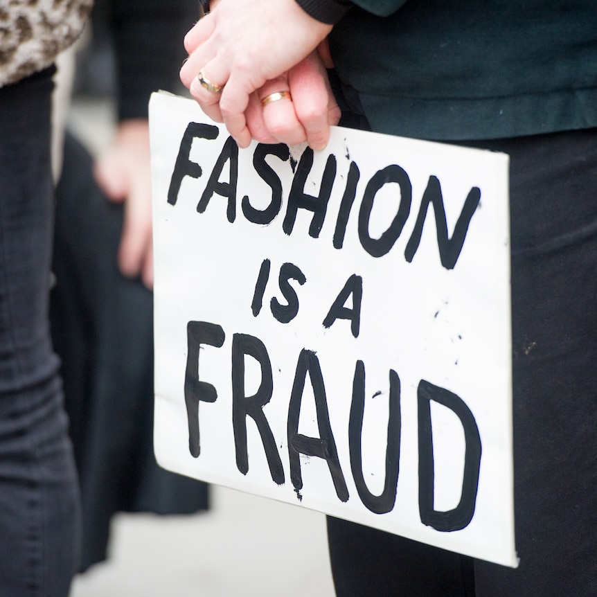 Hands hold a sign that reads 'FASHION IS A FRAUD'.