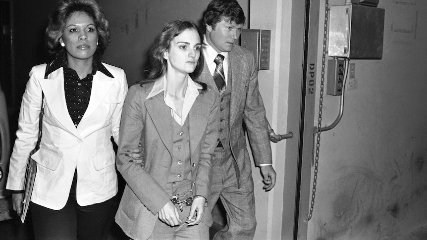 A young woman in a three-piece suit and handcuffs is escorted by a man and a woman