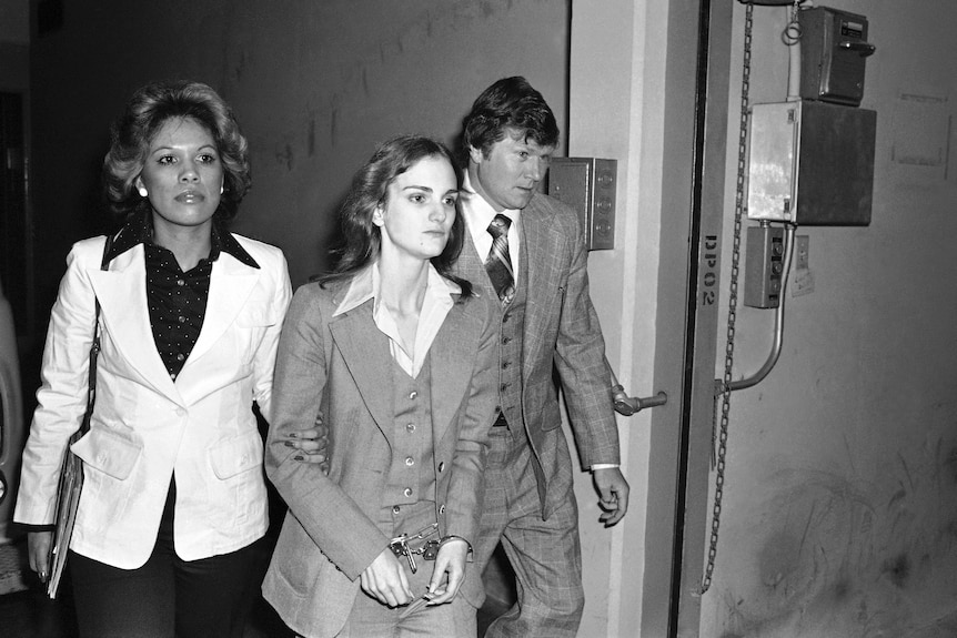 A young woman in a three-piece suit and handcuffs is escorted by a man and a woman